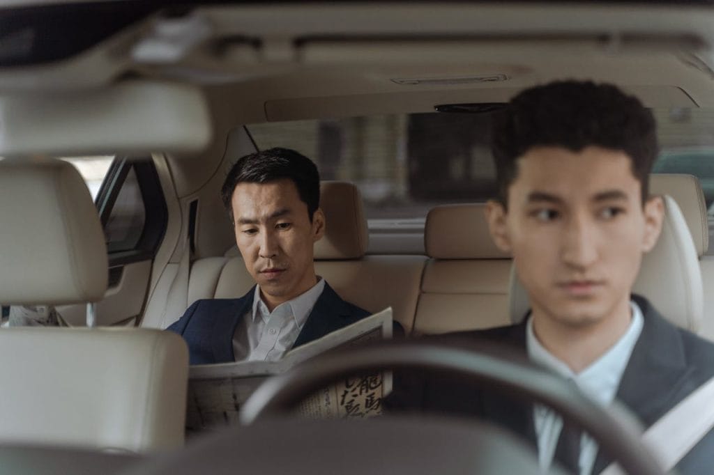 2-men-in-white-suit-sitting-inside-car-9520199 Elegant Asian businessman reading a newspaper inside a luxury car. Professional driver in focus.